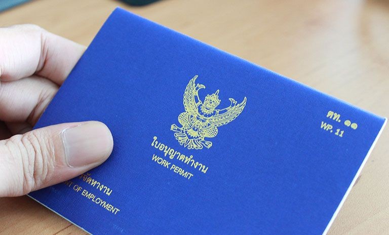 Hand holding a blue Thai work permit booklet with gold emblem and text on a wooden table.